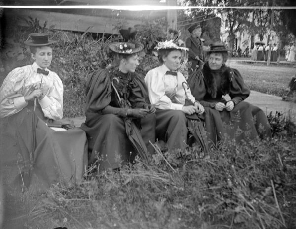 Outdoor portrait of four unidentified women posing sitting on a bench on a lawn. Three women are holding umbrellas. The third woman from the right is identified as the older Poright. A boy is on a sidewalk in the background. In the far background is another group of women standing.