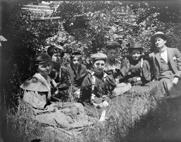 Outdoor portrait of an unidentified group of young men and women posing sitting on the ground in front of tall bushes.
