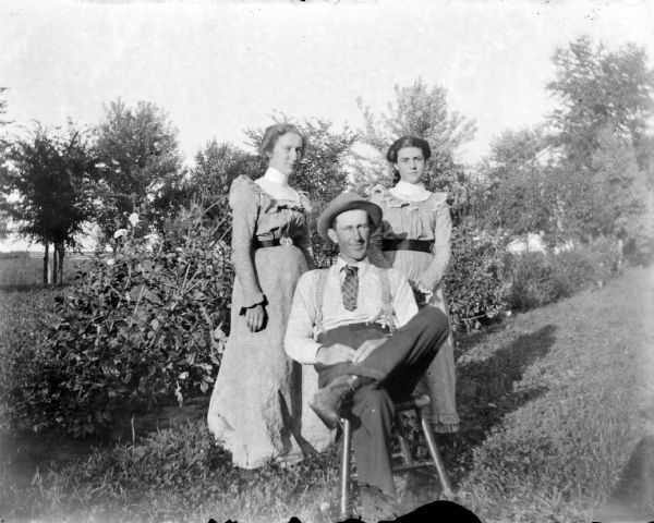 Outdoor group portrait of unidentified man with two women. The man is posing sitting in a chair, in front of and between two women posing standing. They are in a yard with tall bushes and trees.