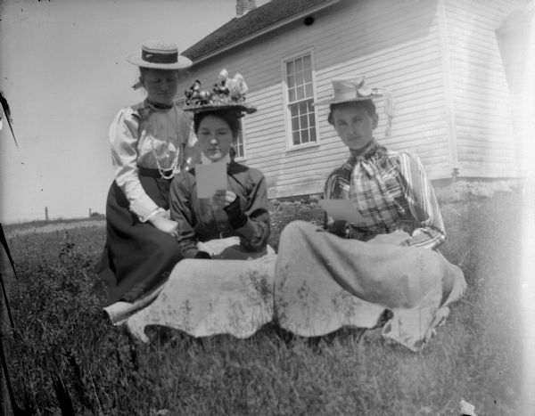 Outdoor group portrait of three unidentified women sitting together on the grass. In the background is a wooden building. The woman on the left is posing kneeling and is wearing a dark-colored skirt and light-colored blouse and a straw hat. The woman in the center, and the woman on the right are posing sitting, and are reading pieces of paper they are holding in their hands. They are both wearing light-colored skirts, blouses and straw hats.