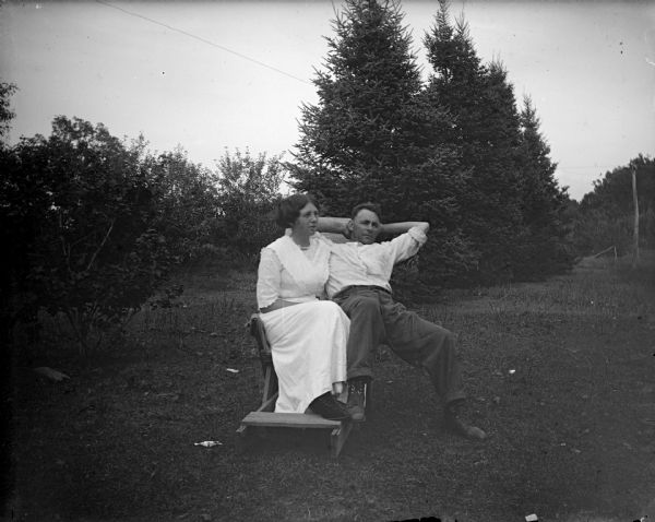 Outdoor portrait of an unidentified woman and man posing sitting in a chair or sled in a grassy yard in front of trees and bushes. The woman is wearing a light-colored dress, and the man, who is resting his head in his arms, is wearing dark-colored trousers and a light-colored shirt.