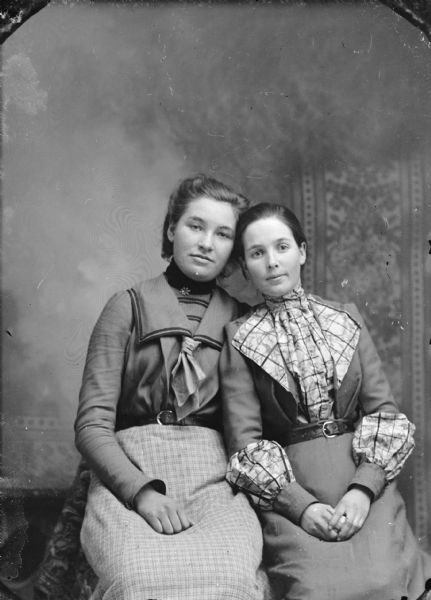 Three-quarter length studio portrait in front of a painted backdrop of two unidentified women posing sitting and leaning against each other. They are both wearing light-colored skirts, belts and blouses, but of different styles.