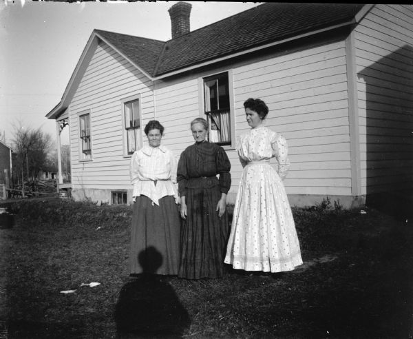 Exterior portrait of a three unidentified women posing standing in a yard. The woman on the left is wearing a dark-colored skirt and light-colored blouse. The older woman in the center is wearing a dark-colored dress. The woman on the right is wearing a light-colored dress. In the background a girl is inside the house looking out the window. The shadow of the photographer is in the foreground.