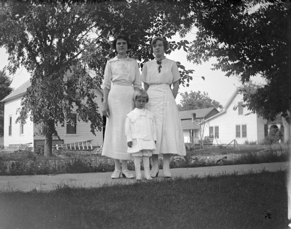 Outdoor group portrait of a child posing standing between two women posing standing on a wooden walkway. Across the street are trees and wooden buildings. The women are wearing light-colored dresses, and the child is wearing light shirt trousers and a blouse.