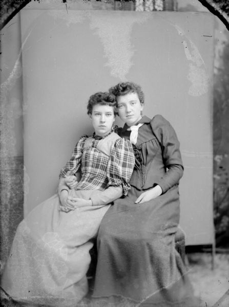 Studio portrait in front of a painted backdrop of two young unidentified women posing sitting. The woman on the left is leaning on the woman on the right. The woman on the left is wearing a light-colored skirt and plaid blouse, and the woman on the right is wearing a dark-colored dress with a necktie.