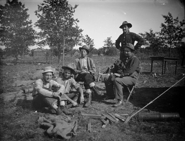 Outdoor group portrait of a five European American men posing sitting and standing in a yard. Identified from left to right as unidentified man, unidentified man, Albert McDonald, Louis Gocheidel, and Fred Werner. Louis is holding a dog on his lap.