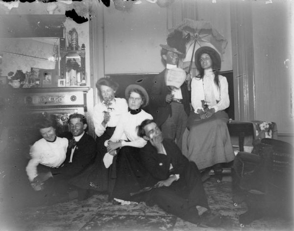 Indoor group portrait of six unidentified people. There are four women and two men posing in the corner of a room. The man on the right is standing and holding an umbrella and a hand fan.