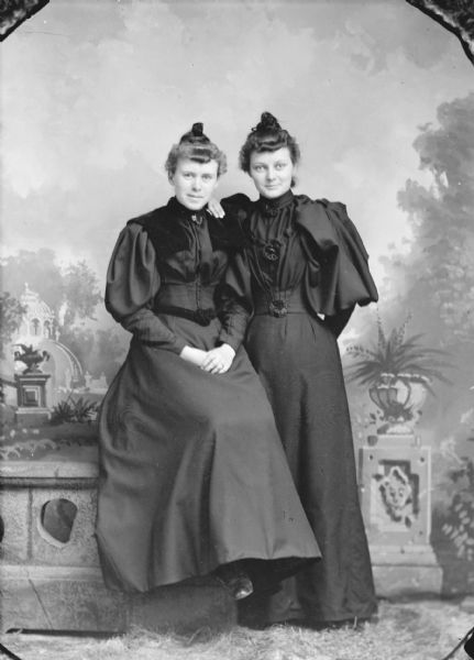 Studio portrait in front of a painted backdrop of two unidentified women. The woman on the left is posing sitting on a prop stone wall, and the woman on the right is standing. They are both wearing dark-colored dresses.