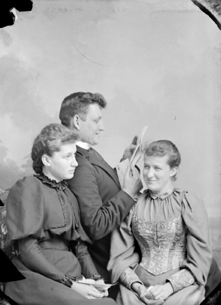 Studio portrait in front of a painted backdrop of three unidentified people posing sitting. The man in the center is wearing a suit jacket and reading a letter. The woman on the left is wearing a dark-colored dress, and the woman on the right is wearing a light-colored dress.