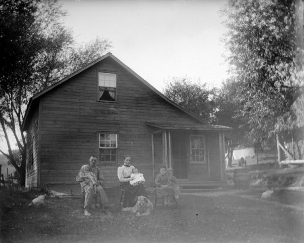 Outdoor group portrait of an unidentified family group, including a man, woman, girl, and infant posing sitting in the yard in front of a wooden house with a dog. Chickens are pecking around in the yard.