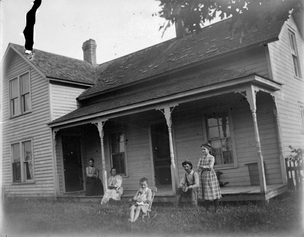 Outdoor group portrait of an unidentified family group posing standing and sitting in the yard and on the porch of a wooden house.