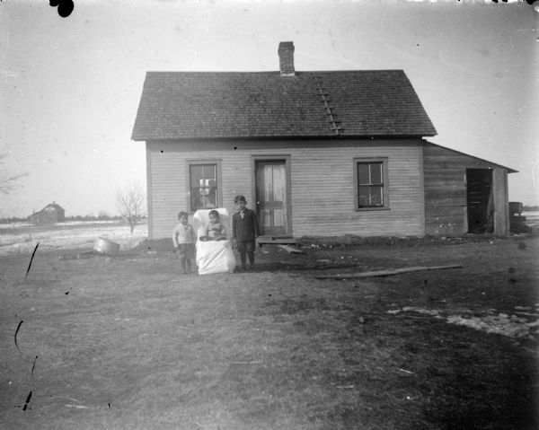 Outdoor group portrait of three unidentified children posing in the yard in front of a small wooden house. Two children are standing on either side of a younger child who is sitting in a chair covered with a white cloth.