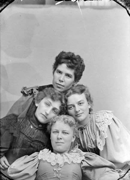 Studio group portrait of four unidentified women posing sitting with their heads close together. The woman on the left is wearing a dark-colored dress, and the other women are wearing lighter-colored dresses. One of the women is identified as perhaps Sadie Dimmick Nolop.