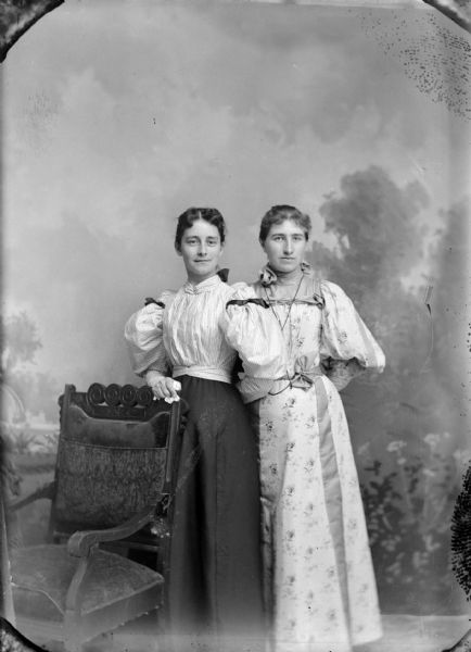 Studio portrait in front of a painted backdrop of two women posing standing. The woman on the left is identified as Flora LeClair, and she is wearing a dark-colored skirt and light-colored blouse and has her hand on the back of a chair. The woman on the right, unidentified, is wearing a light-colored dress.