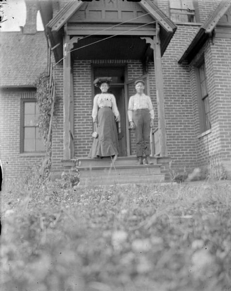 Outdoor portrait of an unidentified man and woman posing standing on the wooden porch of a brick building. The vantage point of the camera is from the ground. Identified as the home on the northwest corner of Jefferson and Water streets, the Best residence.