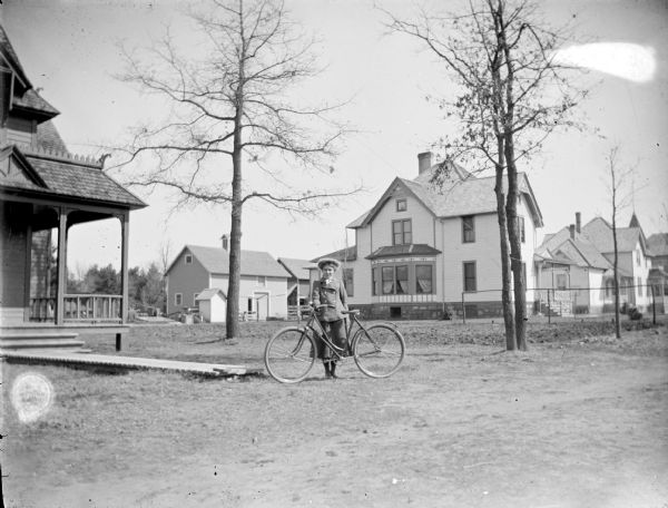 Outdoor portrait of an unidentified woman posing standing with a bicycle in front of several wooden buildings. The street is identified as Marsh Street, and the house directly behind the woman and slightly to the left is identified as the Marsh residence. The house on the far right is identified as the Lochen residence.