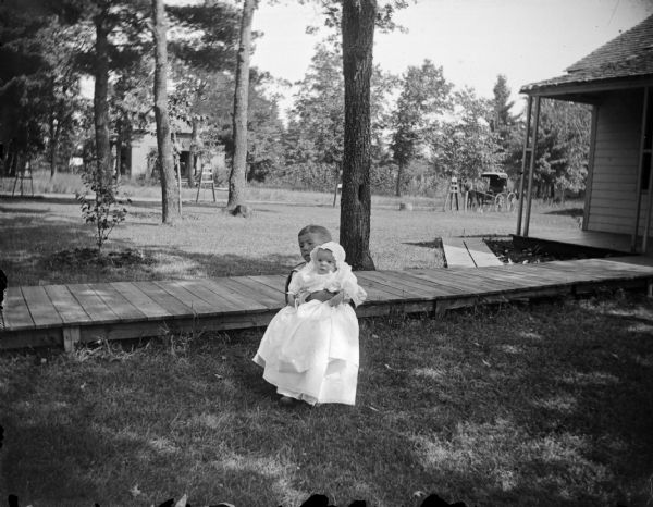 Outdoor portrait of an unidentified child sitting in a chair and holding an infant in their lap near a wooden walkway leading to a house on the right. Across the yard in the background is a horse and carriage. Residence identified as the residence of R.A. Jones and Carol Kinley at the corner of Fourth and Madison Streets.