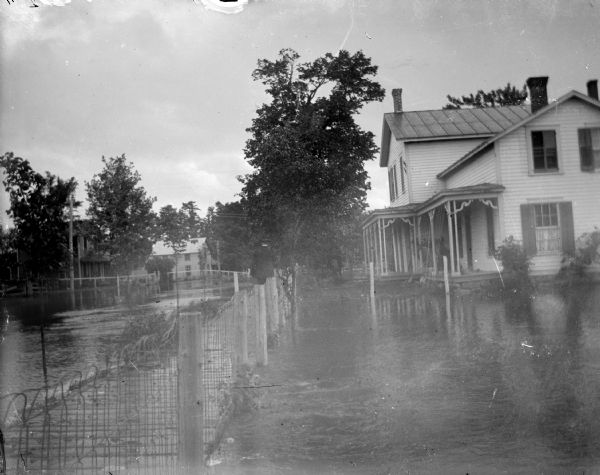 View along fence line towards a wooden house and wooden walkway. Identified as 227 South Street, built in 1868 by J. Chandler, and later owned by W. Taylor. The street and lawn are flooded. A man is walking down the walkway towards the house.