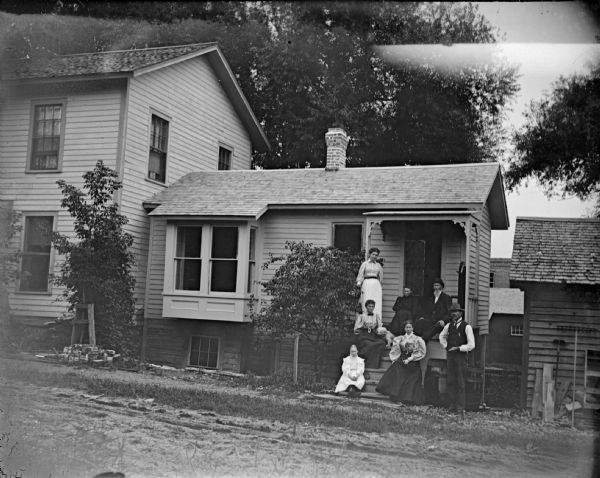 Outdoor group portrait from across street of a European American family posing standing and sitting on the porch of a small wooden house. The house is identified as the residence of L. Dimmick, later Nigara, at 356 Main Street, on the southeast corner of Main and Fourth Streets.
