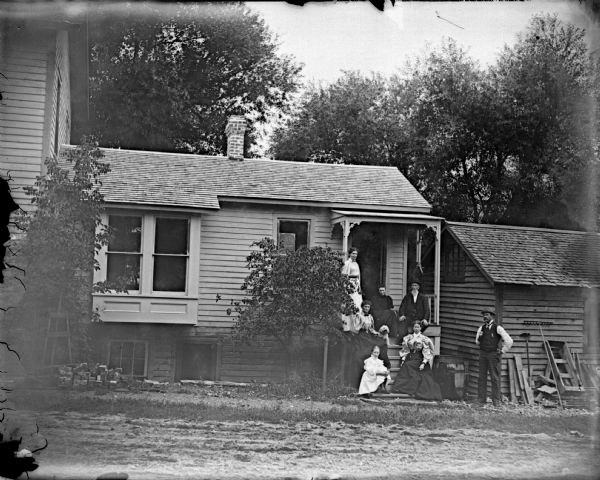 Outdoor group portrait of a European American family posing standing and sitting on the porch of a small wooden house. The house is identified as the residence of L. Dimmick, later Nigara, at 356 Main Street, on the southeast corner of Main and Fourth Streets.