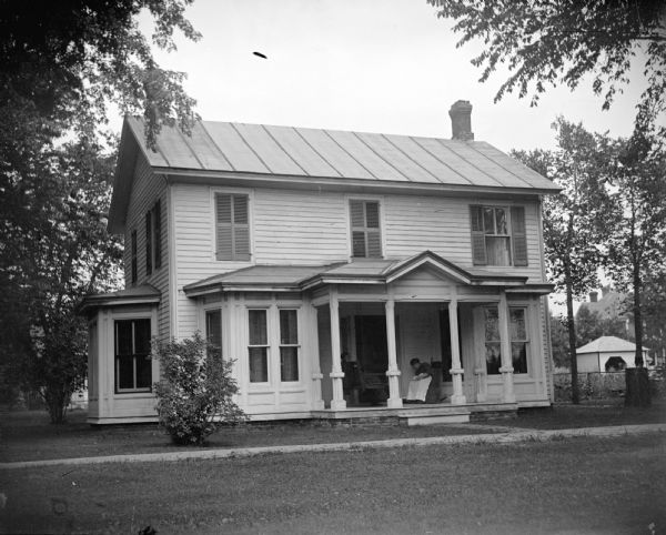 View across lawn towards a European American woman posing sitting on the porch of a wooden house. The house is identified as the residence of Dr. Darnell Gaillardent, later the home of Sullivan and Tom Mills, at 514 Harrison Street, on the northeast corner of Harrison and Fifth Streets. The house was moved to the middle of the block, just west of the Spaulding House in 1950 when the Methodist Church was built.