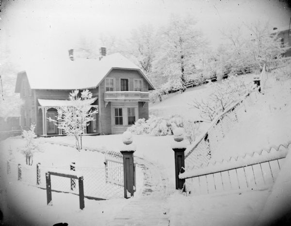 View from street towards a fence surrounding the yard of a house near a hill. The ground is covered with snow. The house is identified as the residence of Dr. Robie on the southwest corner of Main and South Fourth Streets.