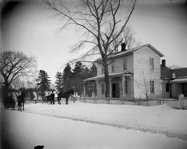 View across street of a two horse-drawn sleighs on the street in front of a two-story wooden house, surrounded by snow. The house is identified as the residence of Johnny Johnson, at the southwest corner of South Second and Johnson Streets.