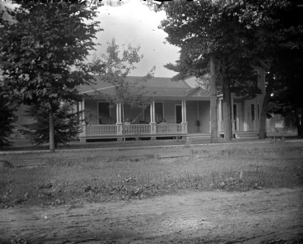 View across street towards two individuals posing sitting on the porch of a house. The house is identified as the residence of the Oderbolz, Pauquette, and Bowman families, northwest corner of Fifth and Van Buren Streets.
