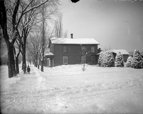 View down sidewalk towards two children posing standing in the snow with a sled near a two-story house. The house is identified as the Hull residence on the northwest corner of Tyler and Ninth Streets.