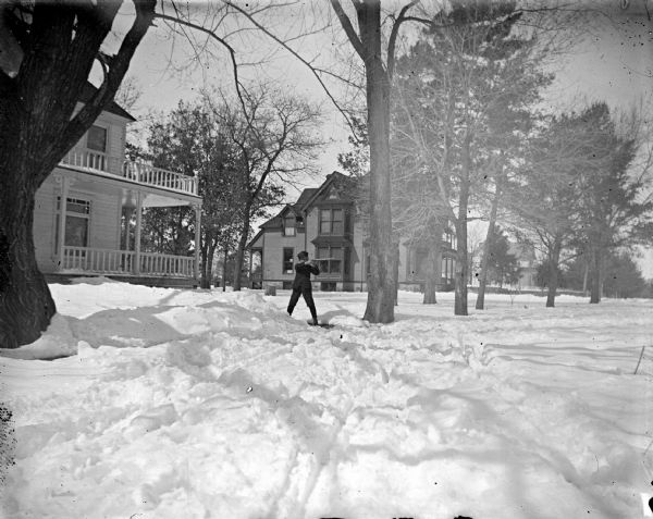 View of a European American boy posing standing and shooting a rifle on the snow-covered ground in the yard in front of a house. The house on the left is identified as the residence of W.H. Richards, and the house on in the background is identified as the residence of L.C. Jones. Both houses are located on North Fourth Street between Harrison and Van Buren Streets.
