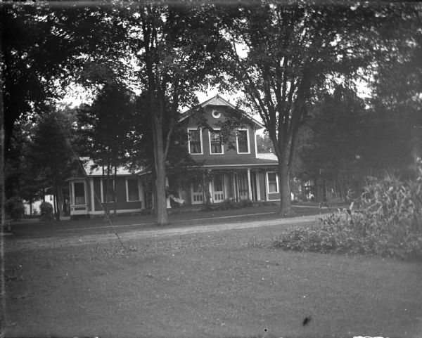 View across lawn towards a two-story wooden house in a yard with several trees in front. The house is identified as the residence of the Pope family on the northwest corner of Sixth and Main Streets.