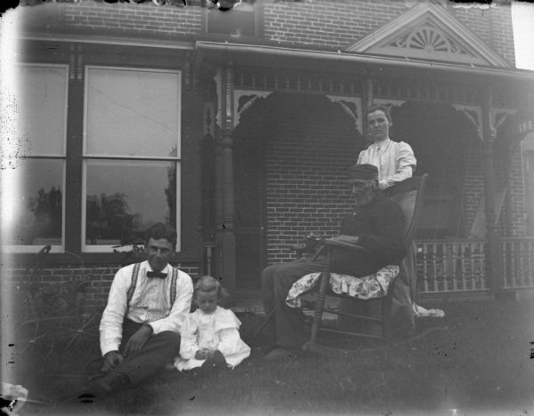 Outdoor group portrait of a European American family, including an elderly man posing sitting in a rocking chair, a woman posing standing, and a man and girl posing sitting on the ground, in front of a brick house. The house is identified as the residence of the Oderboltz family located on the northwest corner of South Third and Pierce Streets.