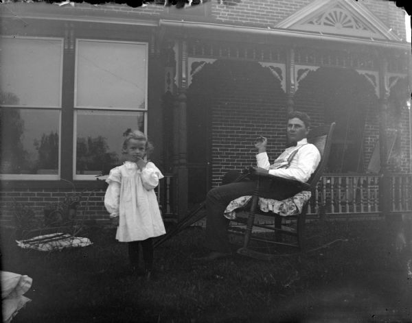 Outdoor view of a European American man posing sitting in a rocking chair and holding crutches. There is a girl posing standing on the left. Behind them is the front of a brick house. House identified as the residence of the Oderboltz family located on the northwest corner of South Third and Pierce Streets.