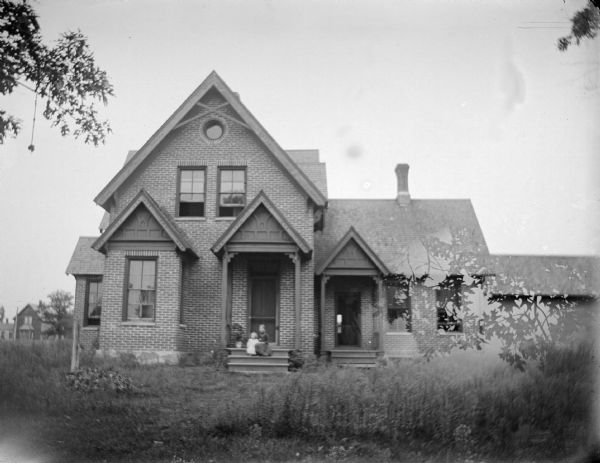 View across yard towards two children posing sitting on the stoop of a brick two-story house. House identified as the residence of Fred Best built on the northwest corner of Water and Jefferson Streets, later owned by Moulderhauer, now the Torgenson Funeral Home. Negative double-exposed with another image.