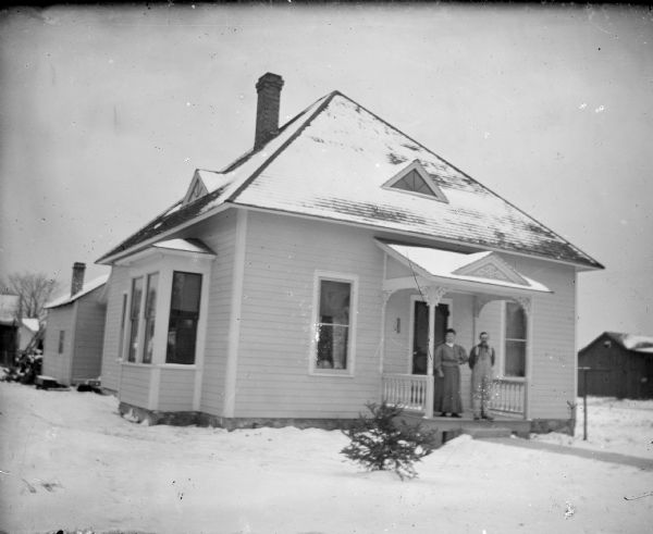 View across snowy yard towards a European American man and woman posing standing on a stoop of a single-story wooden house. Location of house identified as on the south side of Fillmore Street between Sixth and Seventh Streets.