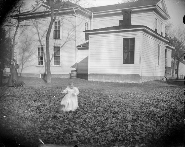 View of a young girl playing in the leaves in the yard of a two-story wooden house. A man and woman and a dog stand watching in the background on the far left. House identified as the residence of W.T. Murray on the southwest corner of Main and Melrose Streets.
