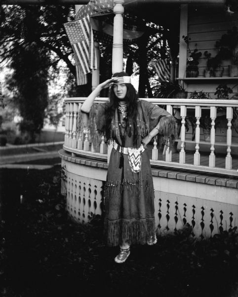 Outdoor portrait of a European American woman posing standing and wearing a Native American costume in front of a wooden porch. The porch is decorated with United States flags and plants. The house is identified as the Maddock's residence.