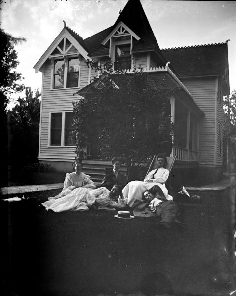 Outdoor group portrait of a European American group. Three women are posing sitting, and a man and woman are posing lying on the ground in front of a two-story wooden house. Another man is lying on the ground behind the women. House identified as the residence of Richenback.
