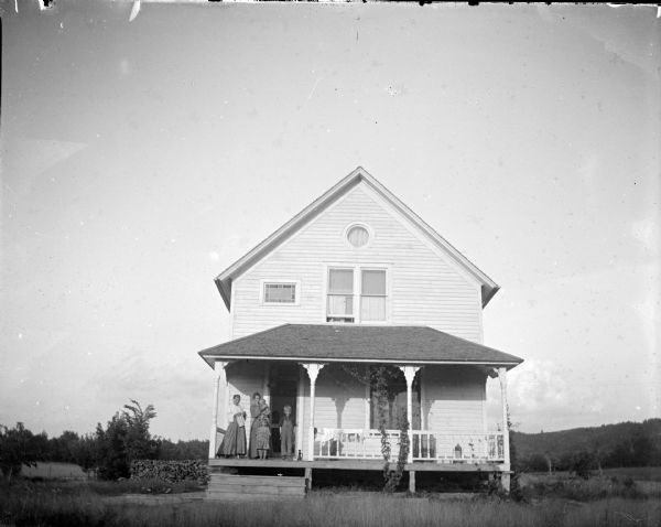 View across yard towards a group of European American people. Two women, two children, and an infant are posing on the porch of a two-story wooden house.