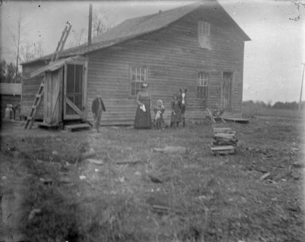 View across yard towards a European American group of people. Standing together in front of the house is a woman, boy, and two girls, with one girl holding the reins of a pony. The building is a small, wooden two-story house with a small roofed entrance.