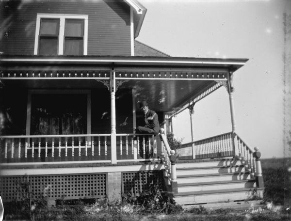 Exterior portrait of a European American man posing sitting on the railing of a porch of a two-story wooden house.