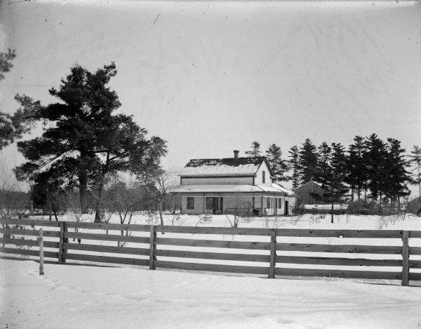 Exterior portrait of a wooden house in the distance surrounded by a large snow-covered yard and wooden fence.
