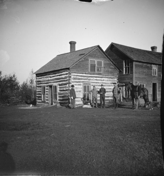 View across yard towards five European American men, two dogs, and a horse posing standing in front of a log cabin. On the right is a wooden board house abutting the cabin.