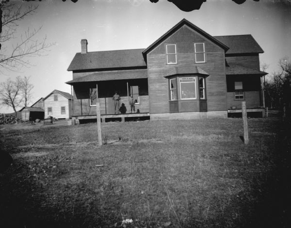 View across lawn of a man and boy posing standing on the porch of a wooden house. There are outbuildings in the background on the left.