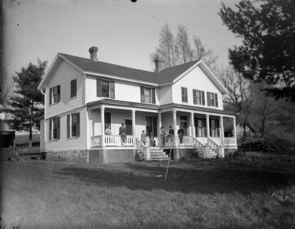 View across yard towards a family group posing sitting and standing on the porch of a large two-story house.
