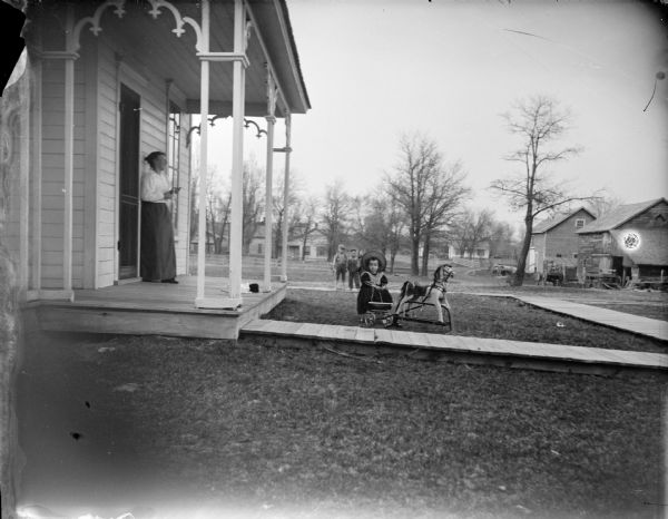 Outdoor view across lawn toward a European American woman posing standing on the porch of a wooden house. A girl is posing sitting on a wagon behind a wooden hobby horse. In the background two small boys are posing standing on a wooden walkway. Houses and outbuildings are in the background.