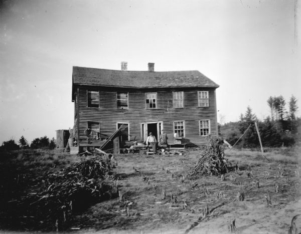 View across yard towards a man and woman posing sitting and standing in front of a dilapidated two-story wooden house with broken windows. In the foreground are stacks of corn stalks.