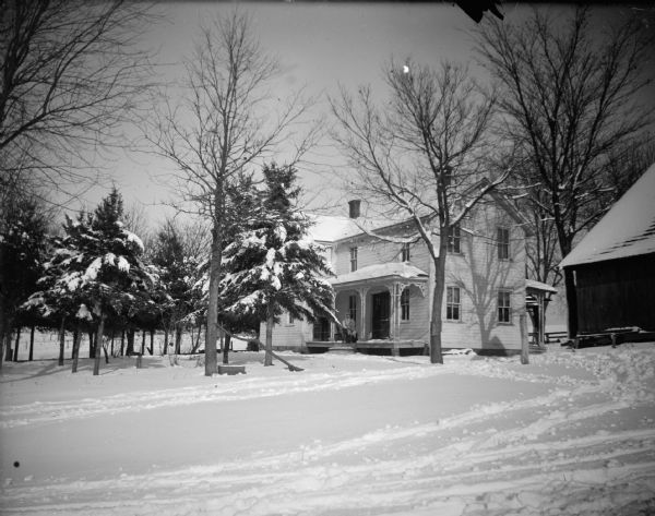 Outdoor view across snow-covered yard towards a woman and child posing while sitting on the porch of a two-story house surrounded by snow-covered trees and yard. Another building is on the right.