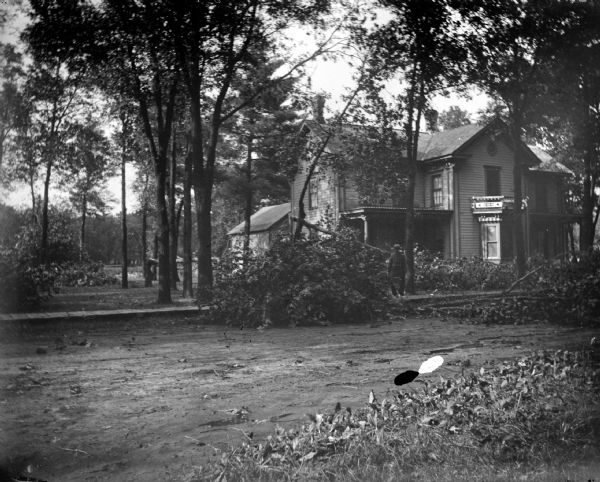 Outdoor view across road towards a man posing standing on a wooden walkway near downed trees and branches and in front of a two-story wooden house.