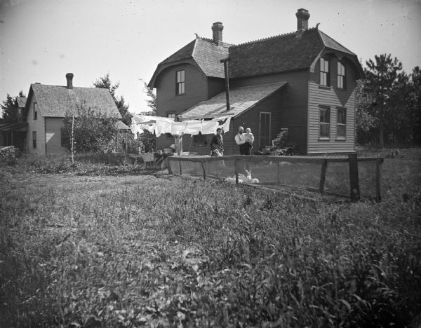 Outdoor view across yard towards a European American family standing together behind a long rabbit hutch in front of laundry hanging on the line. The man is holding a child in his arms, and a woman is standing nearby on the left. They are posing in front of a two-story wooden house.